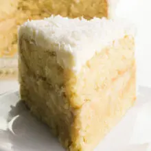 A slice of vegan coconut cake on a plate with the rest of the cake behind it.