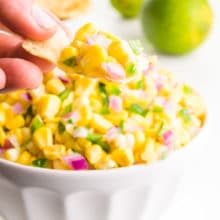 A hand holds a tortilla chip with corn salsa on it. It's sitting above a bowl full of the salsa. There are limes behind it.