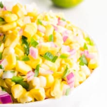 A bowl holds a fresh batch of corn salsa. There's a lime behind it.
