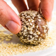A hand holds a date ball, rolling it in sesame seeds.