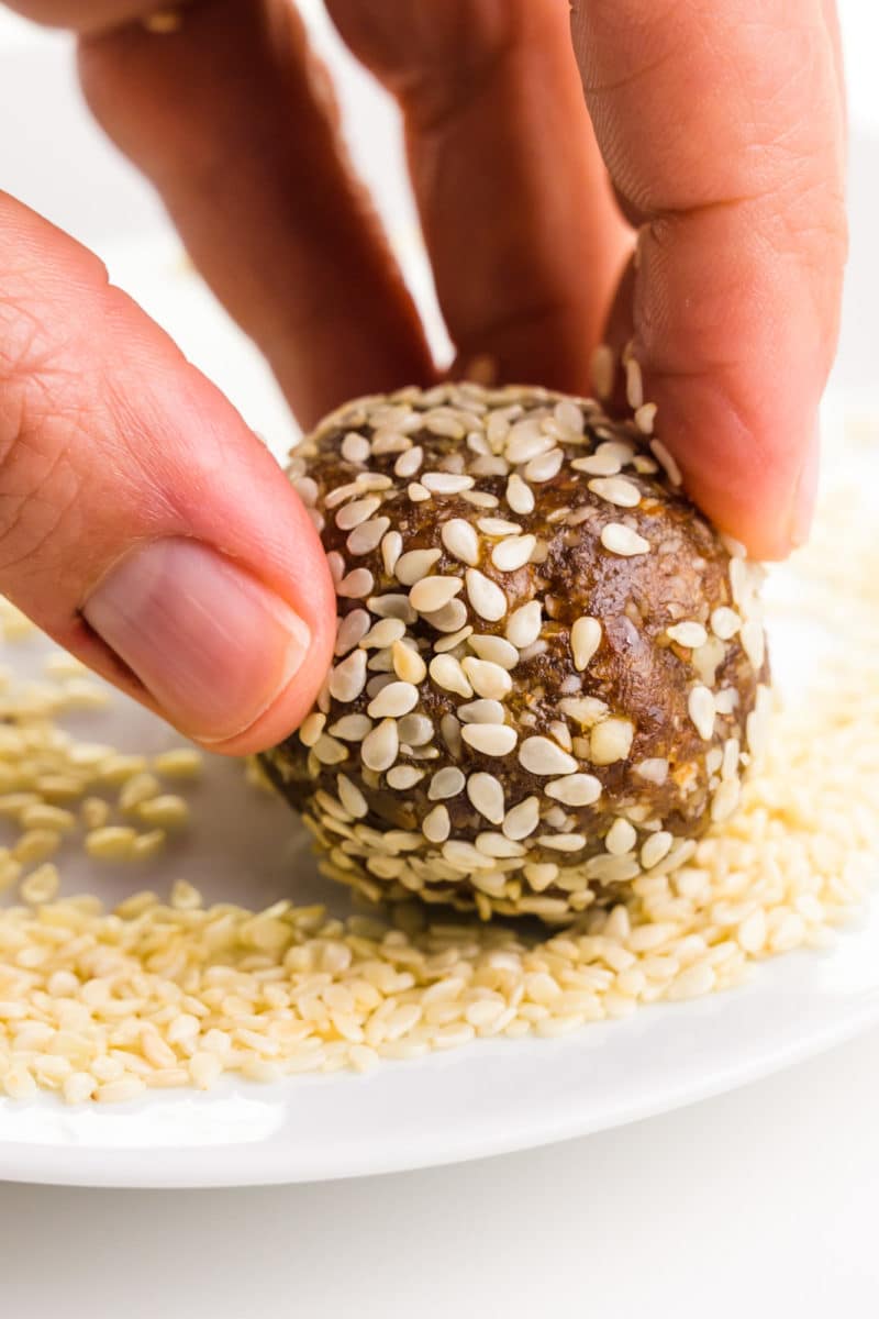A hand holds a date ball, rolling it in sesame seeds.