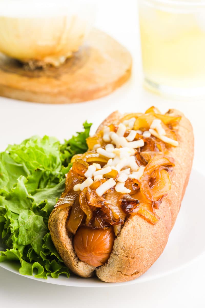 A hot dog is in a bun and is topped with grilled onions and mozzarella cheese. Steamed broccoli is next to the hot dog. there's a cutting board with an onion behind it and a glass of iced tea.