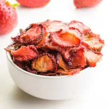 Oven-dried strawberries are in a white bowl, with several fresh strawberries behind it.