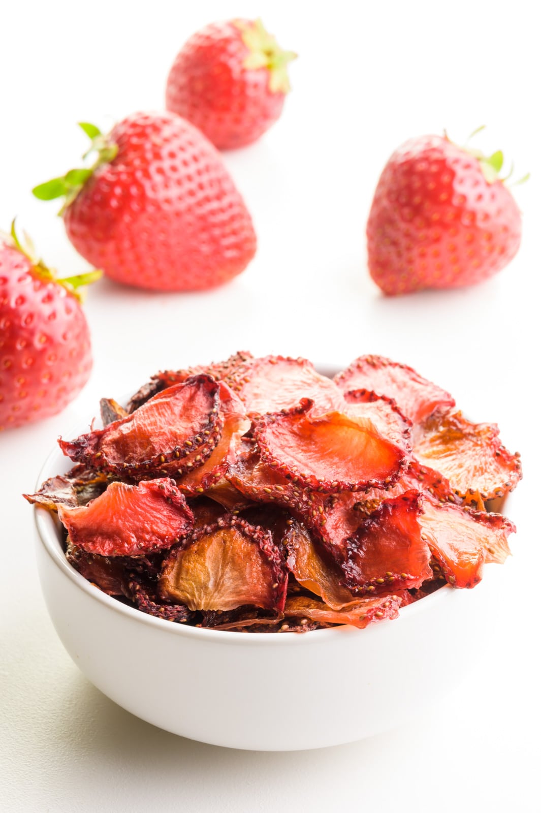 A bowl full of oven-dried strawberries sits in front of fresh strawberries.