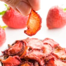 A hand holds a dried strawberry over a bowl of dried strawberry slices. There are fresh strawberries behind the bowl.