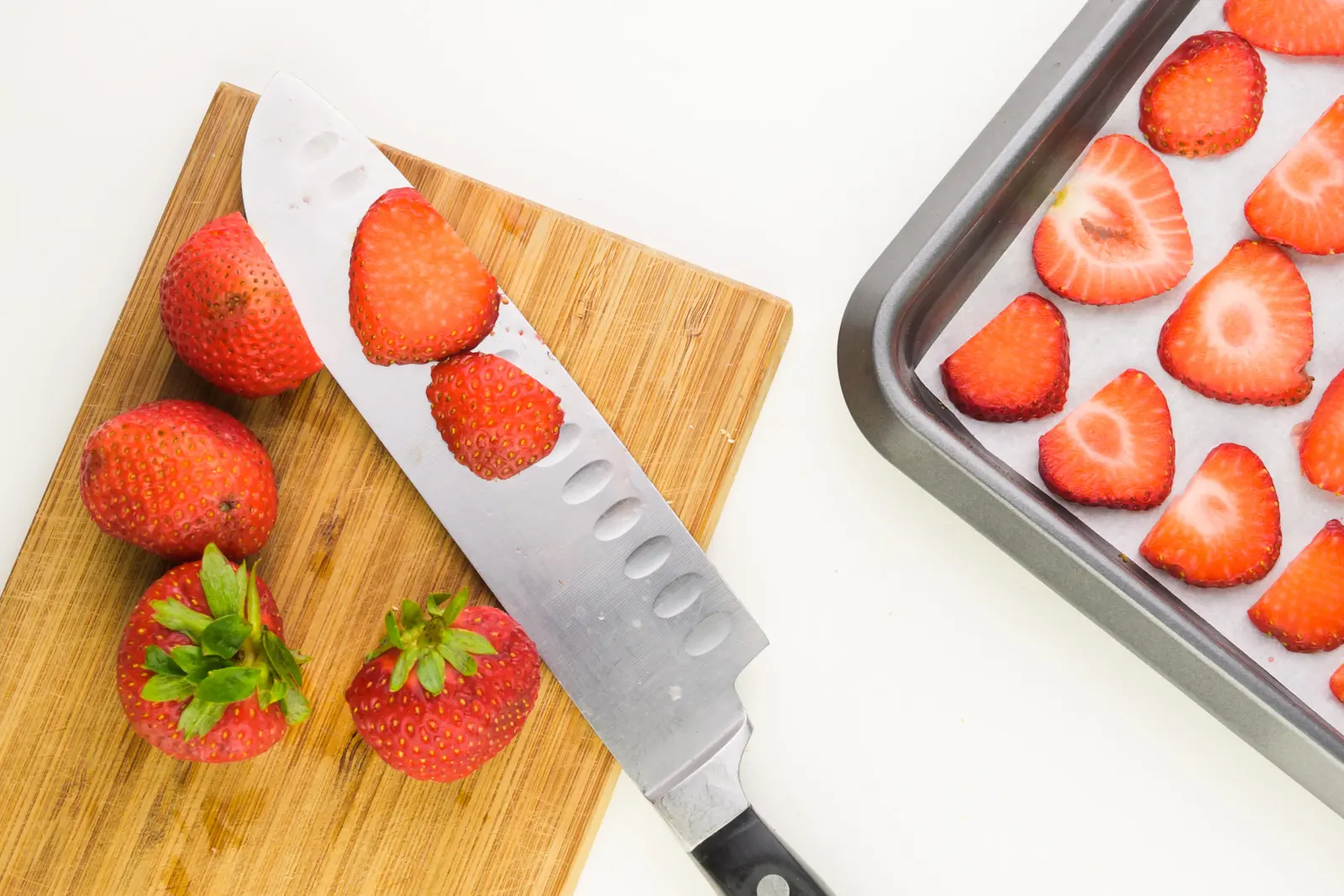 A knife sits on a cutting board with several fresh and sliced strawberries. A pan sits nearby with sliced strawberries in it.