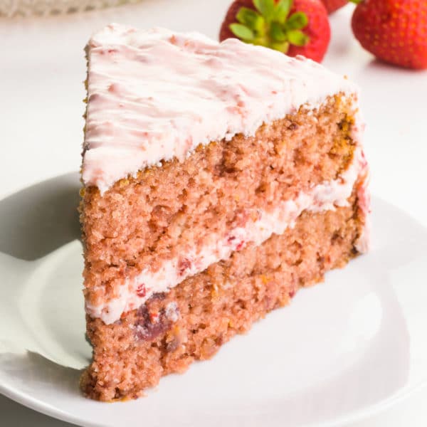 A slice of layered strawberry cake on a plate with fresh strawberries behind it.