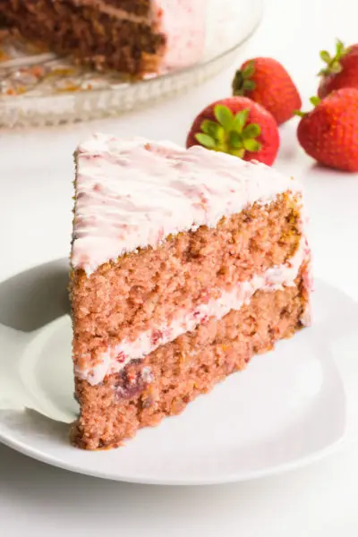 A layered vegan strawberry cake sits on a plate. There are strawberries behind it and the rest of the cake.