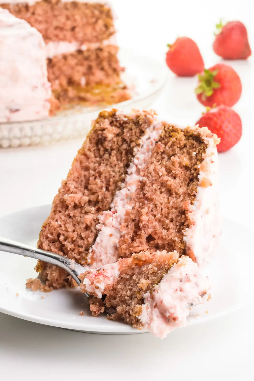 A layered slice of strawberry cake is sitting on a plate with a fork holding a bite. There are fresh strawberries and the rest of the cake behind it.