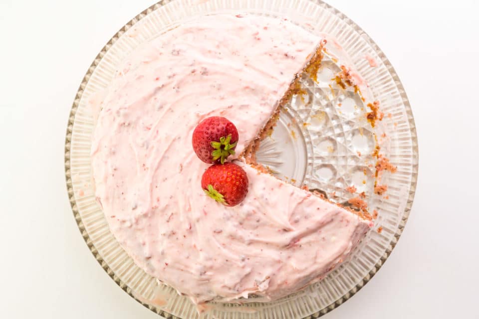 Looking down on a strawberry cake with a slice cut out of it. There are two fresh strawberries on top.