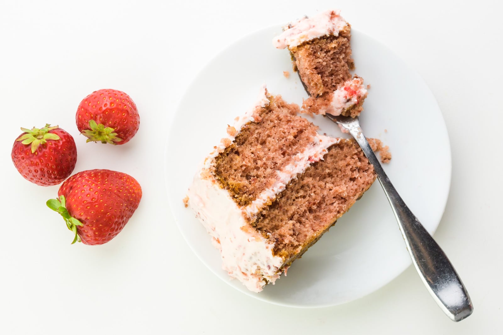 Looking down on a slice of layered strawberry cake with a bite sitting on a fork next to it. There are three fresh strawberries sitting next to it.