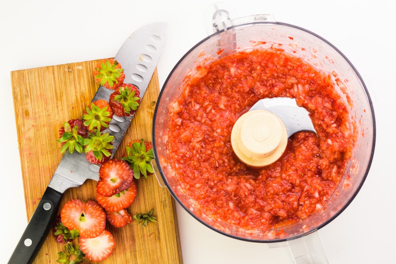 A red sauce is in the bottom of a food processor. There's a knife on a cutting board with freshly cut strawberry bits on the cutting board.