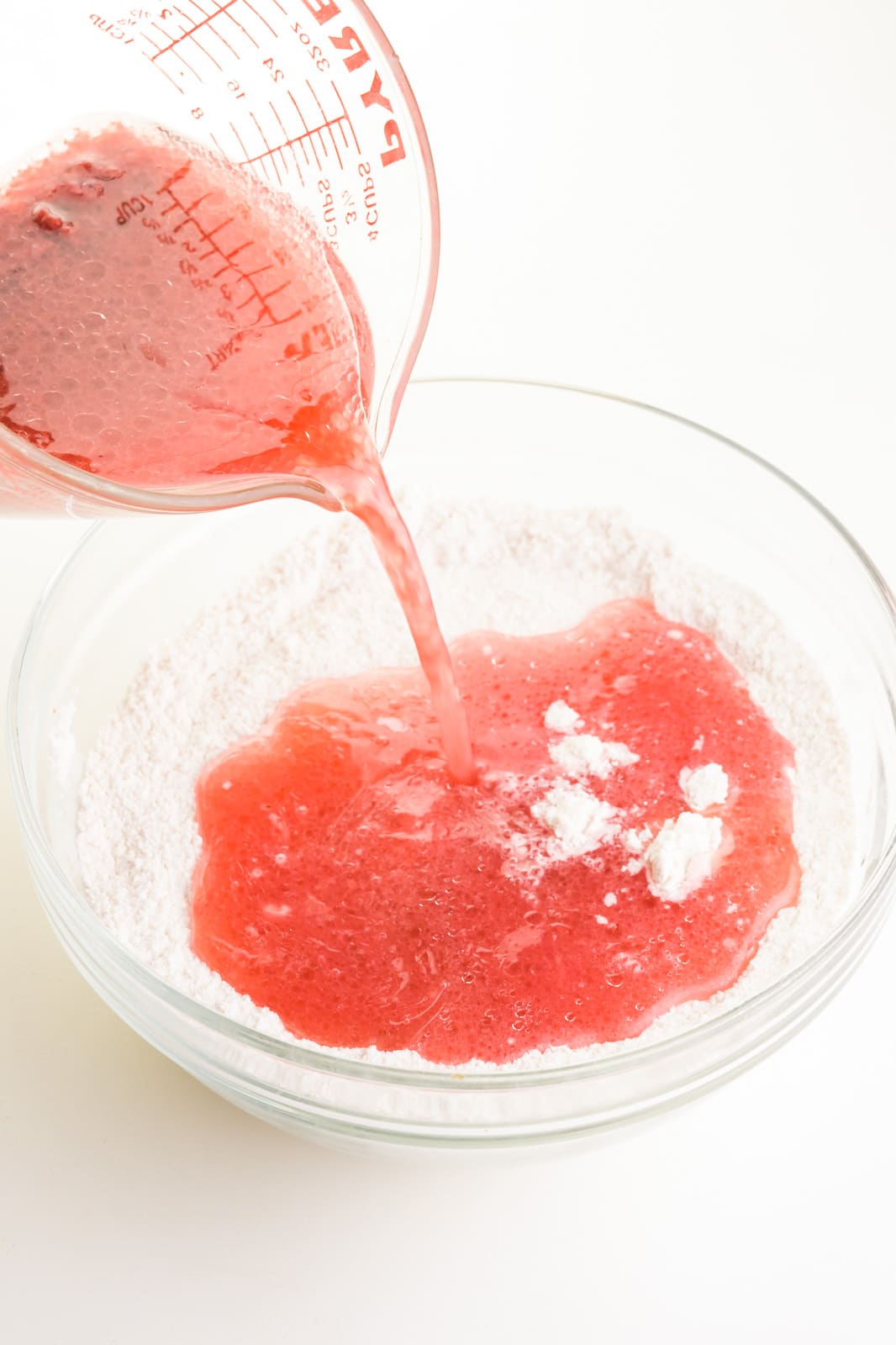 A red liquid is being poured into a glass bowl with a flour mixture. This to make batter for a strawberry cake.