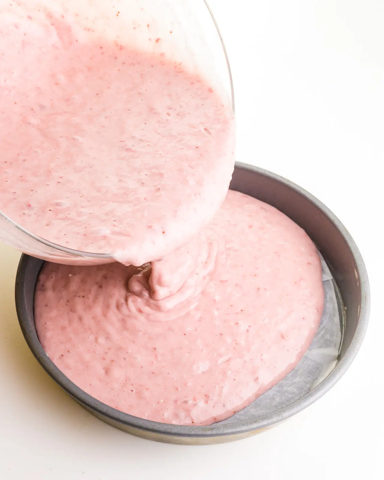 A pink cake batter is being poured into a round baking pan.