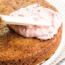 A spatula is spreading strawberry frosting on a round cake.