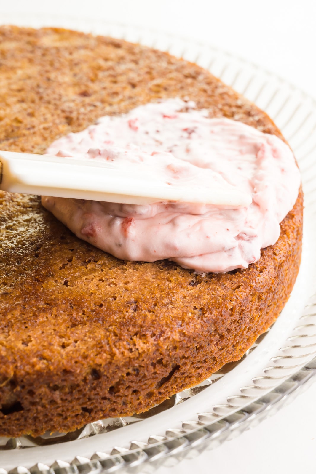 A spatula is spreading strawberry frosting on a round cake.
