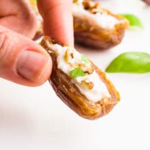 A hand holds a cream cheese stuffed date. There are more dates and basil leaves behind it.