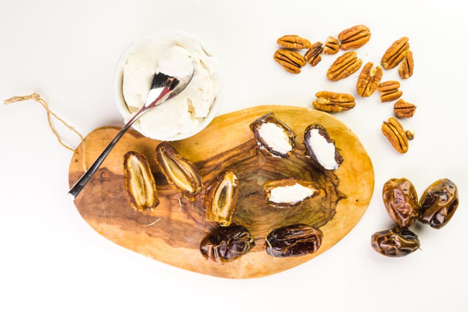 Dates on a cutting board have been cut in half. Some have bee stuffed with vegan cream cheese. A bowl of cream cheese sits next to the board along with several whole dates.
