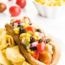 A Tex Mex hotdog is topped with black beans, tomatoes, corn, and more. There are potato chips behind it. Behind this iare some cherry tomatoes and a bowl of corn salsa.