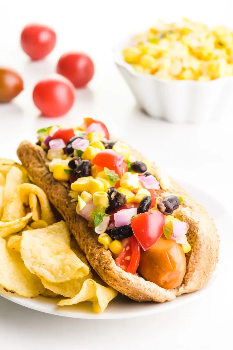 A Tex Mex hotdog is topped with black beans, tomatoes, corn, and more. There are potato chips behind it. Behind this iare some cherry tomatoes and a bowl of corn salsa.