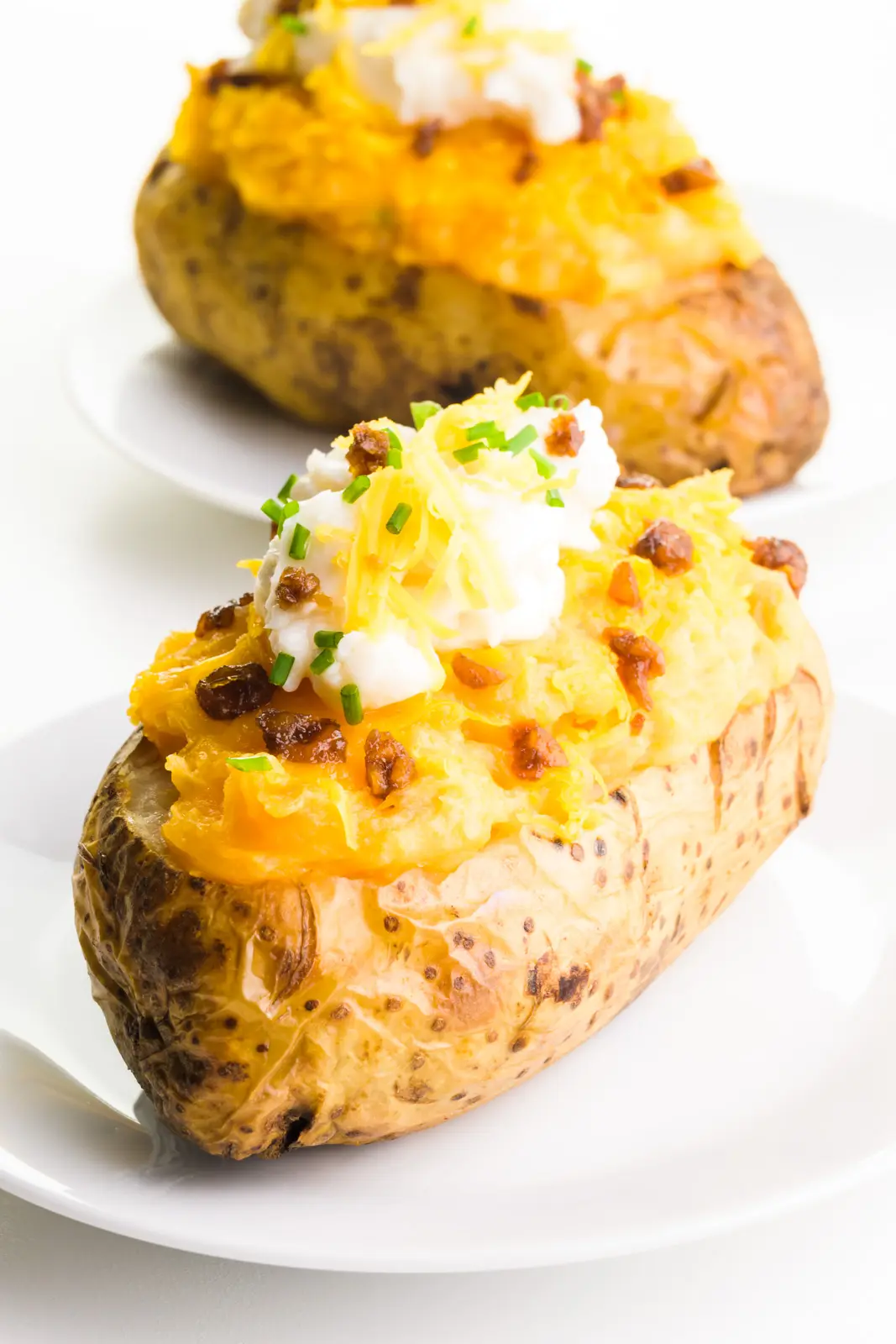 Two vegan twice baked potatoes sit on plates, with vegan bacon, sour cream, and cheese on top.