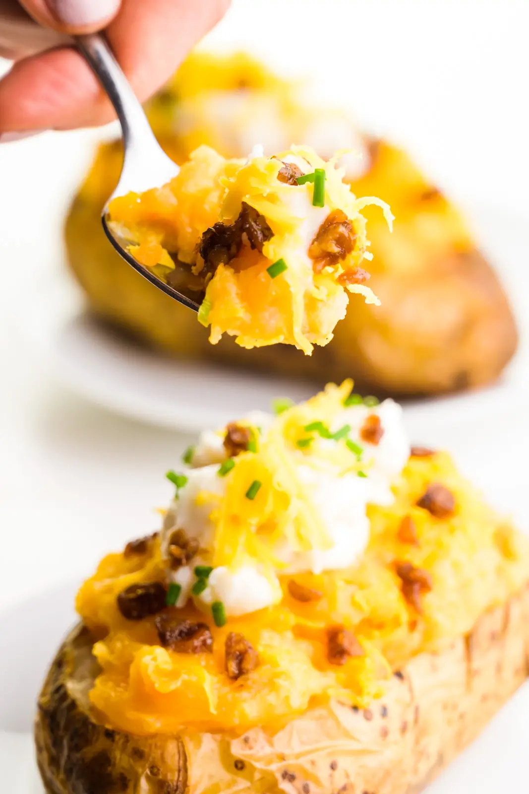 A hand holds a spoon full of potato filling, from vegan twice baked potatoes sitting on a plate behind it.