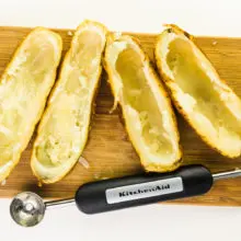 Four baked potatoes have had the centers scooped out. They're sitting on a cutting board next to a melon baller that was used to scoop the potatoes.