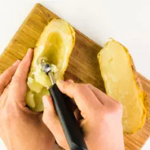 A hand is using a melon baller to scoop the insides of a baked potato that has been cut in half.