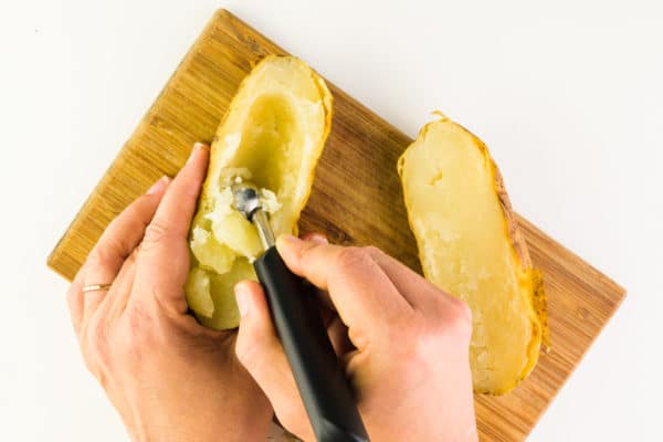 A hand is using a melon baller to scoop the insides of a baked potato that has been cut in half.