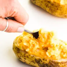 A hand holds a spoon and is placing cheesy mashed potato filling into a scooped out baked potato. Another potato sits behind it.