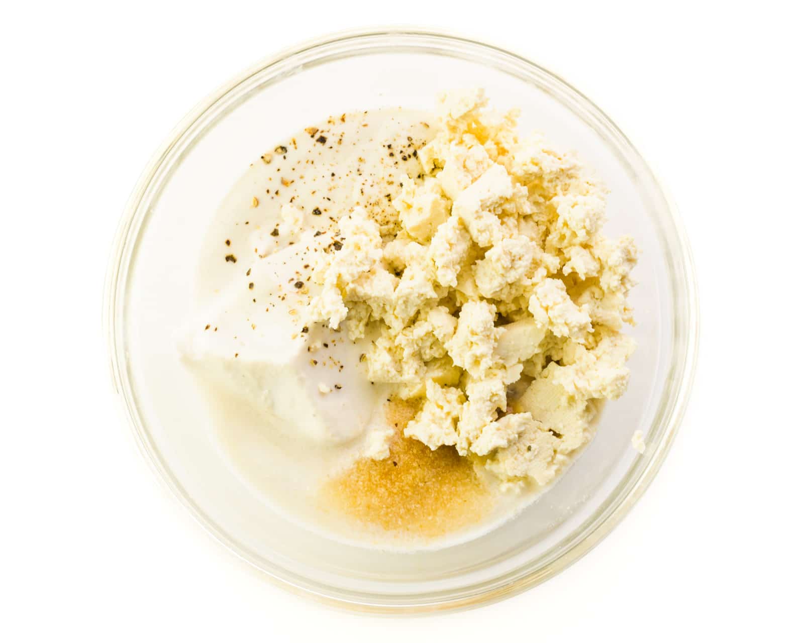 Ingredients are in the bottom of a glass bowl, including vegan sour cream, garlic, powder, and crumbled tofu.