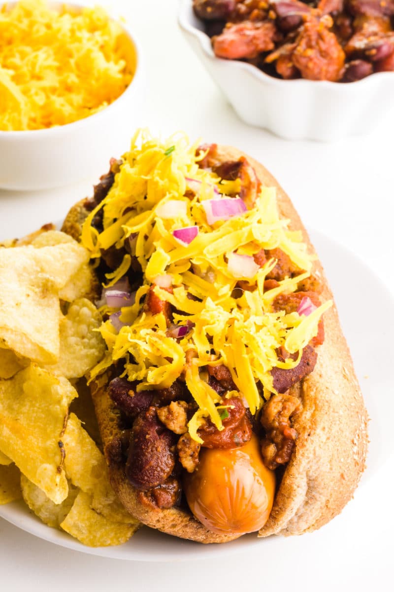 A vegan chili dog has a bowl of cheese and a bowl of chili behind it.
