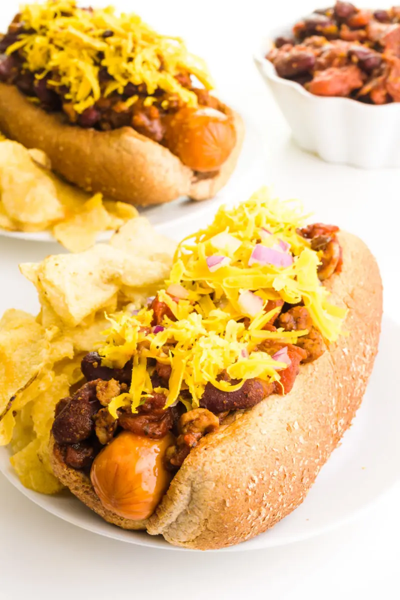 A vegan chili dog has cheese on top. There's another one behind it along with a bowl of chili.