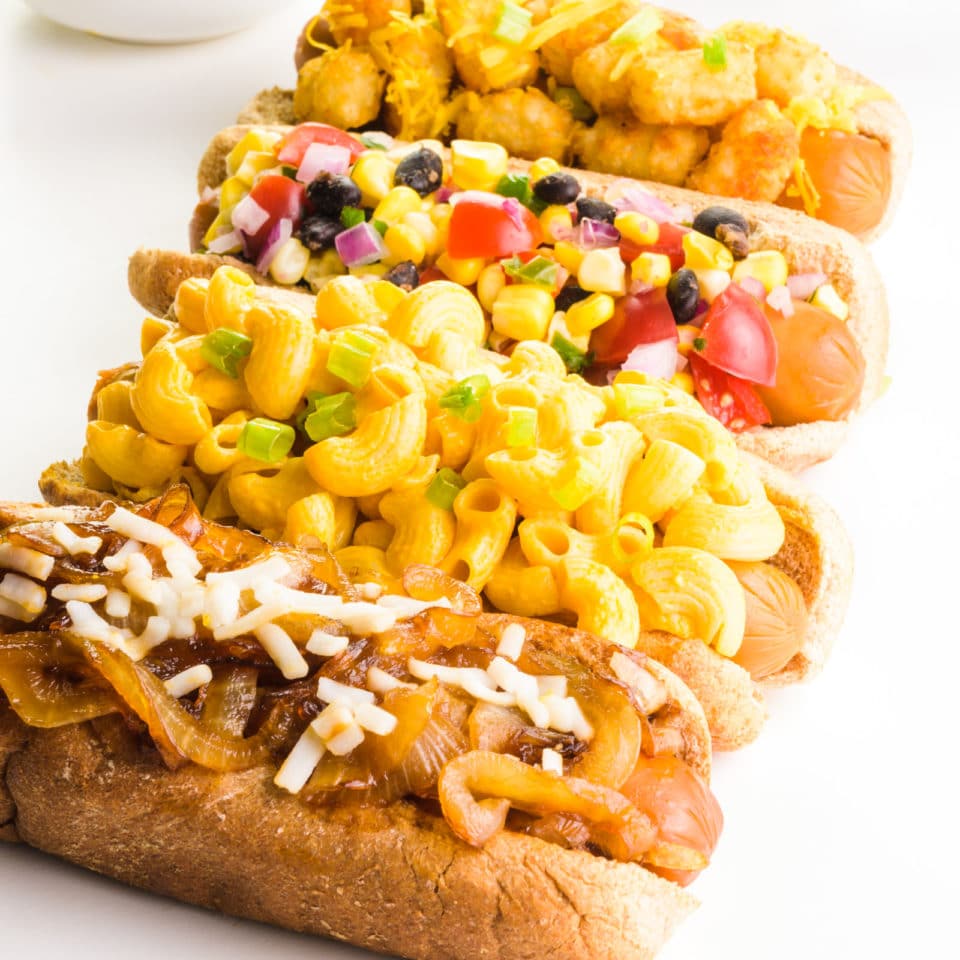 Four vegan hot dogs with different toppings, like mac and cheese, onions, and more, are all lined up. There's a bowl with more toppings behind them.