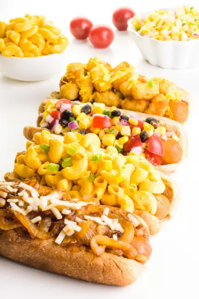Four hotdogs are lined up in a row, They all have different toppings, like onions, tater tots, and more. There are bowls of toppings behind them.