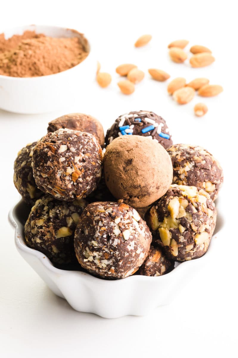 A bowl of chocolate balls all have different toppings and sit in front of a bowl of cocoa powder and nuts in the background.