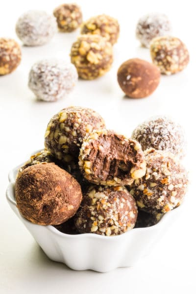 Several vegan truffles are in a bowl with more truffles behind them. They're all coated with different toppings, like chopped nuts and coconut.