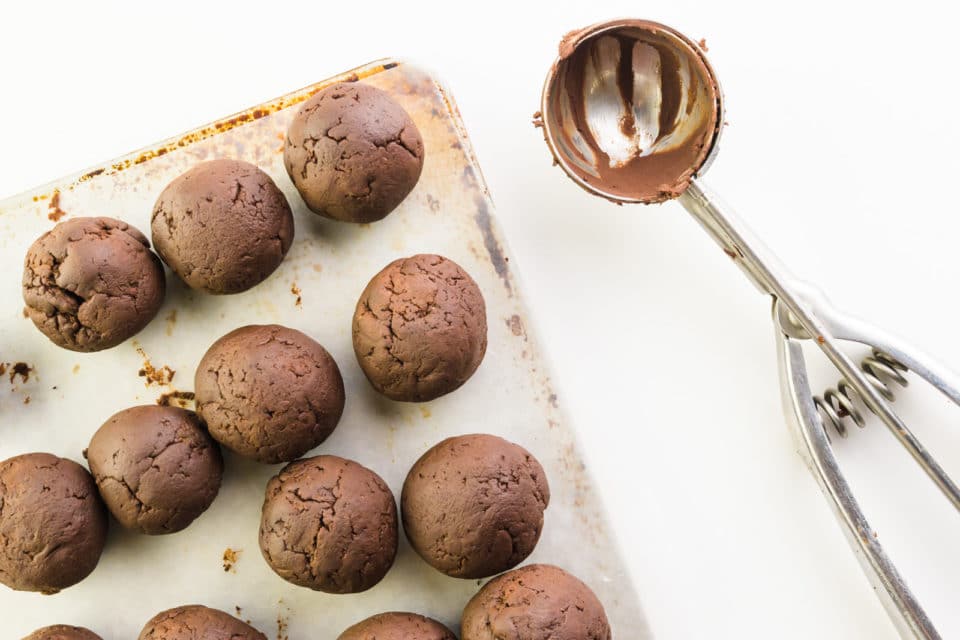 Chocolate balls sit on a pan lined with waxed paper. A cookie scoop with bits of chocolate on it sits next to it.