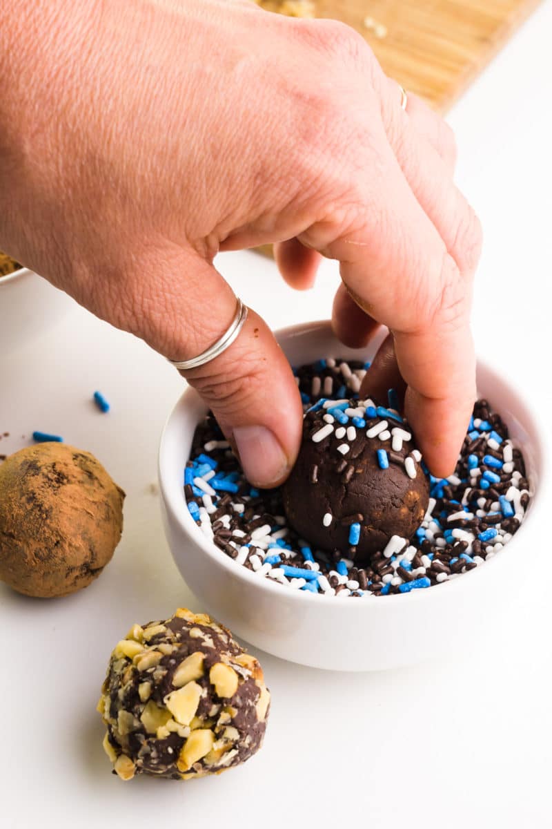 A hand holds a chocolate truffle and is rolling it in a bowl of sprinkles. More truffles sit near the bowl.