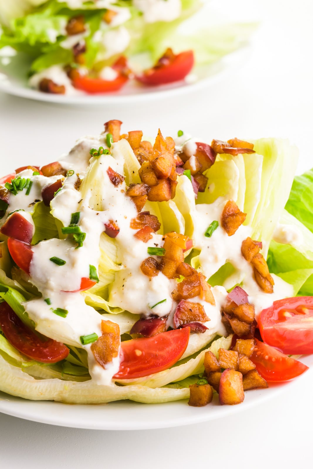 A wedge salad sits on a plate, with chopped tomatoes, smoky apple bits, and vegan dressing on top. There's another salad behind it.