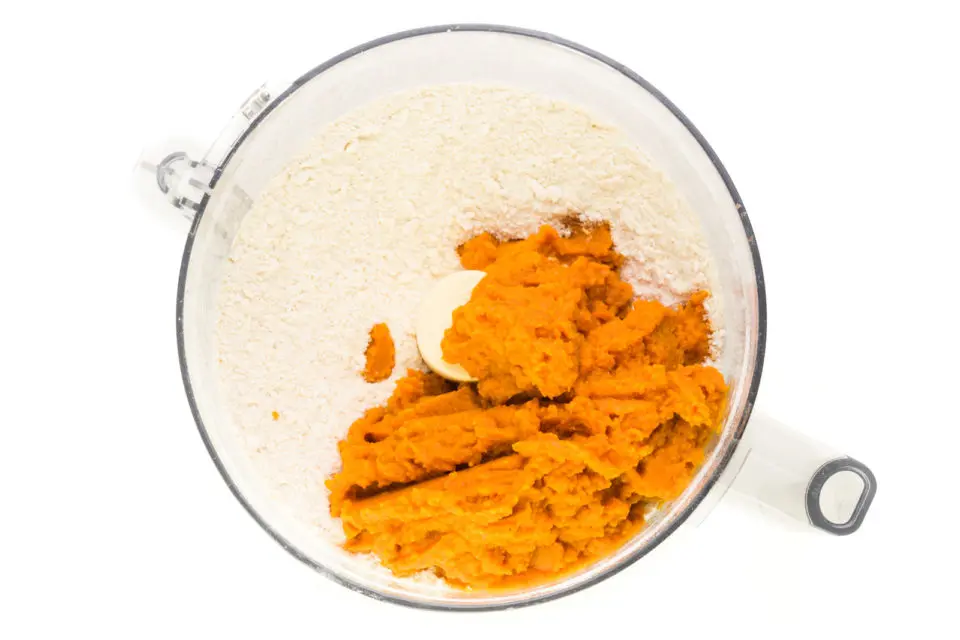 Pureed pumpkin has been added to a food processor bowl along with four ingredients.
