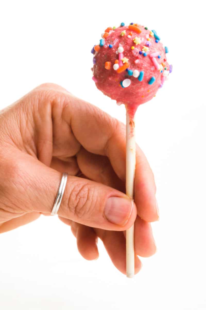 A hand holds a pink cake pop by the stick. The cake pop is pink with colorful sprinkles.