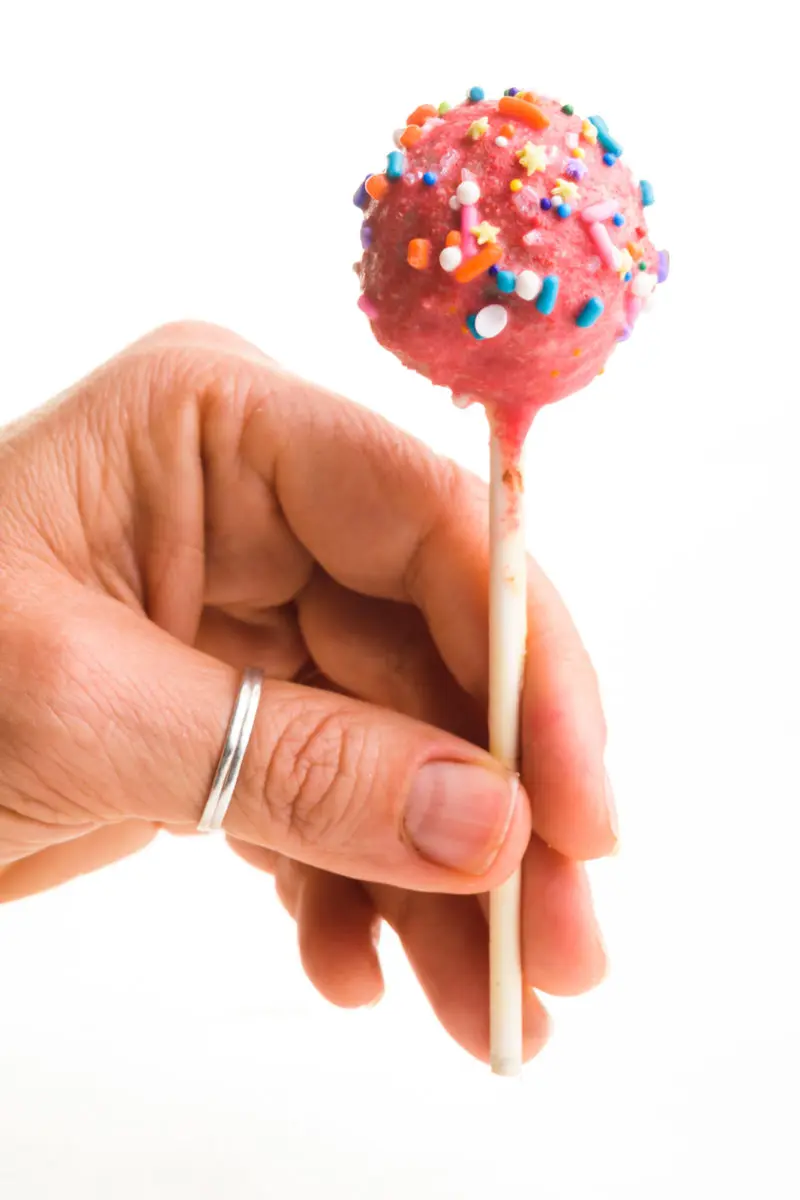 A hand holds a pink cake pop by the stick. The cake pop is pink with colorful sprinkles.