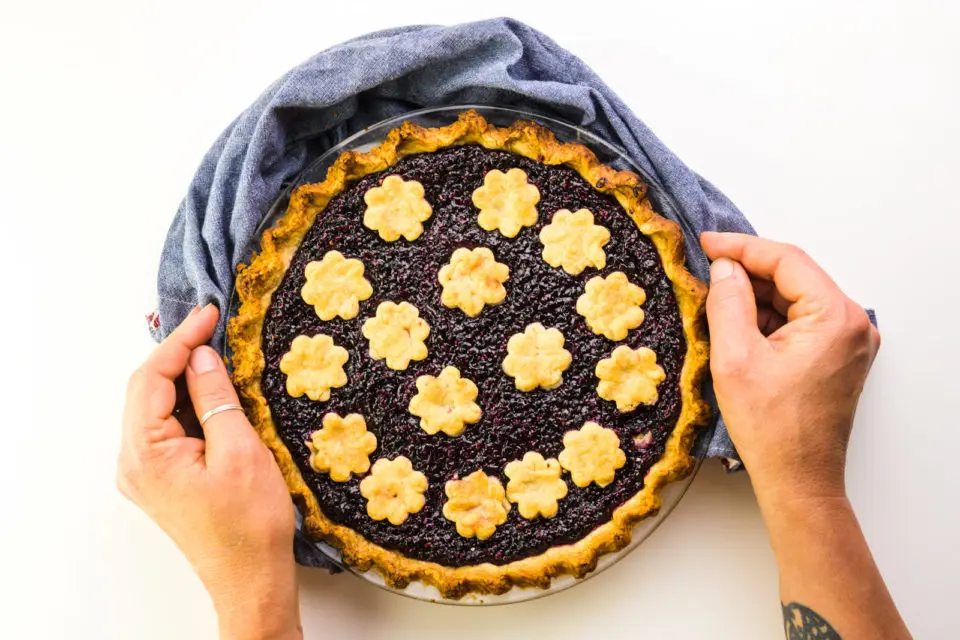 Two hands reach in with a blue kitchen towel, to grab a freshly baked pie.