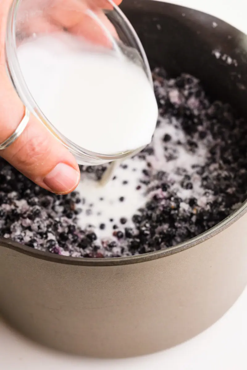 A hand holds a small glass bowl full of a cornstarch slurry, pouring it into a sauce pan full of berries.