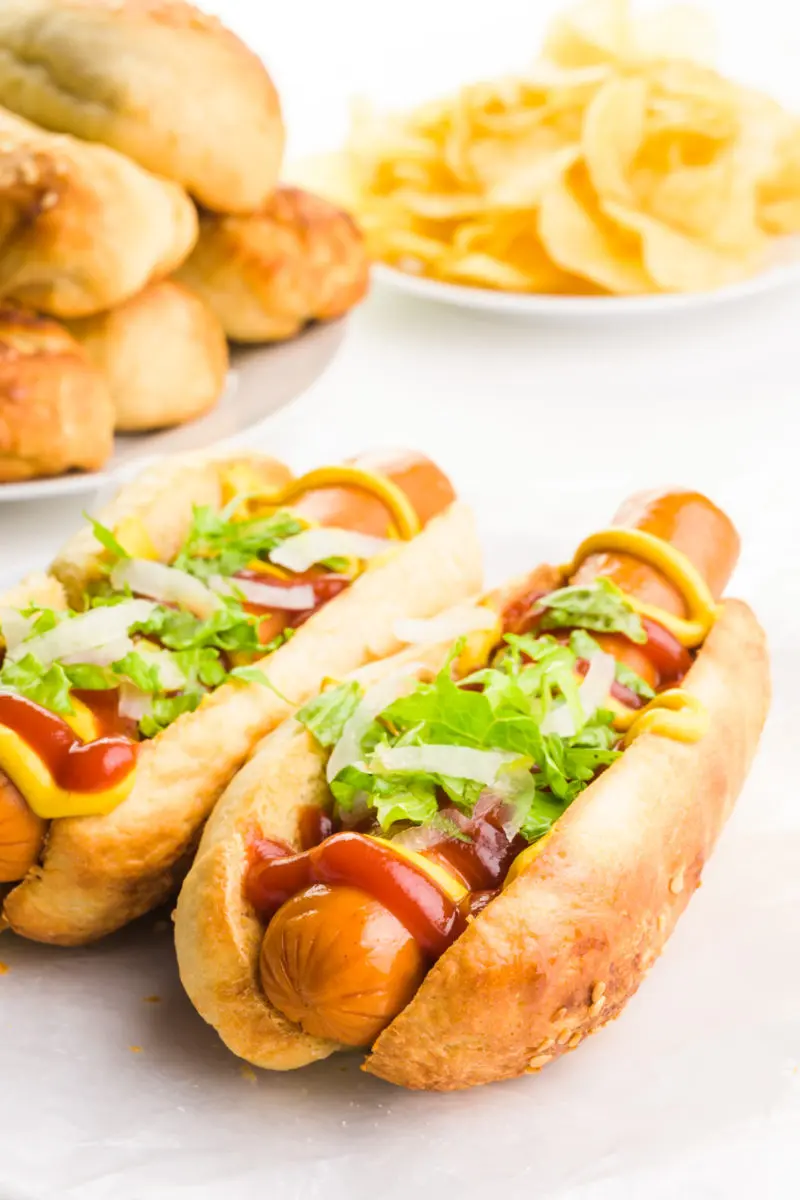 Two hot dog buns have hot dogs inside with lots of toppings, such as ketchup and mustard. There's a plate with more buns behind it and a plate with potato chips.