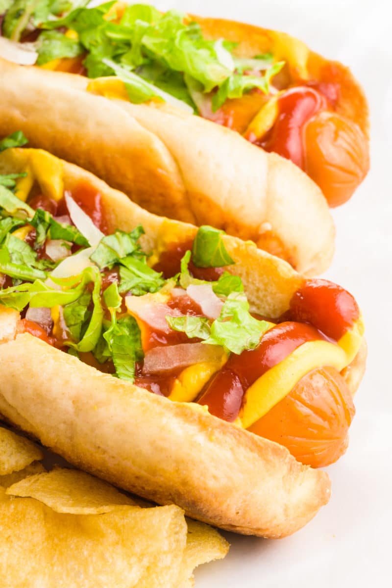 Two hot dog buns are filled with hot dogs topped with ketchup, mustard, greens, and more. There are potato chips on the plate too.