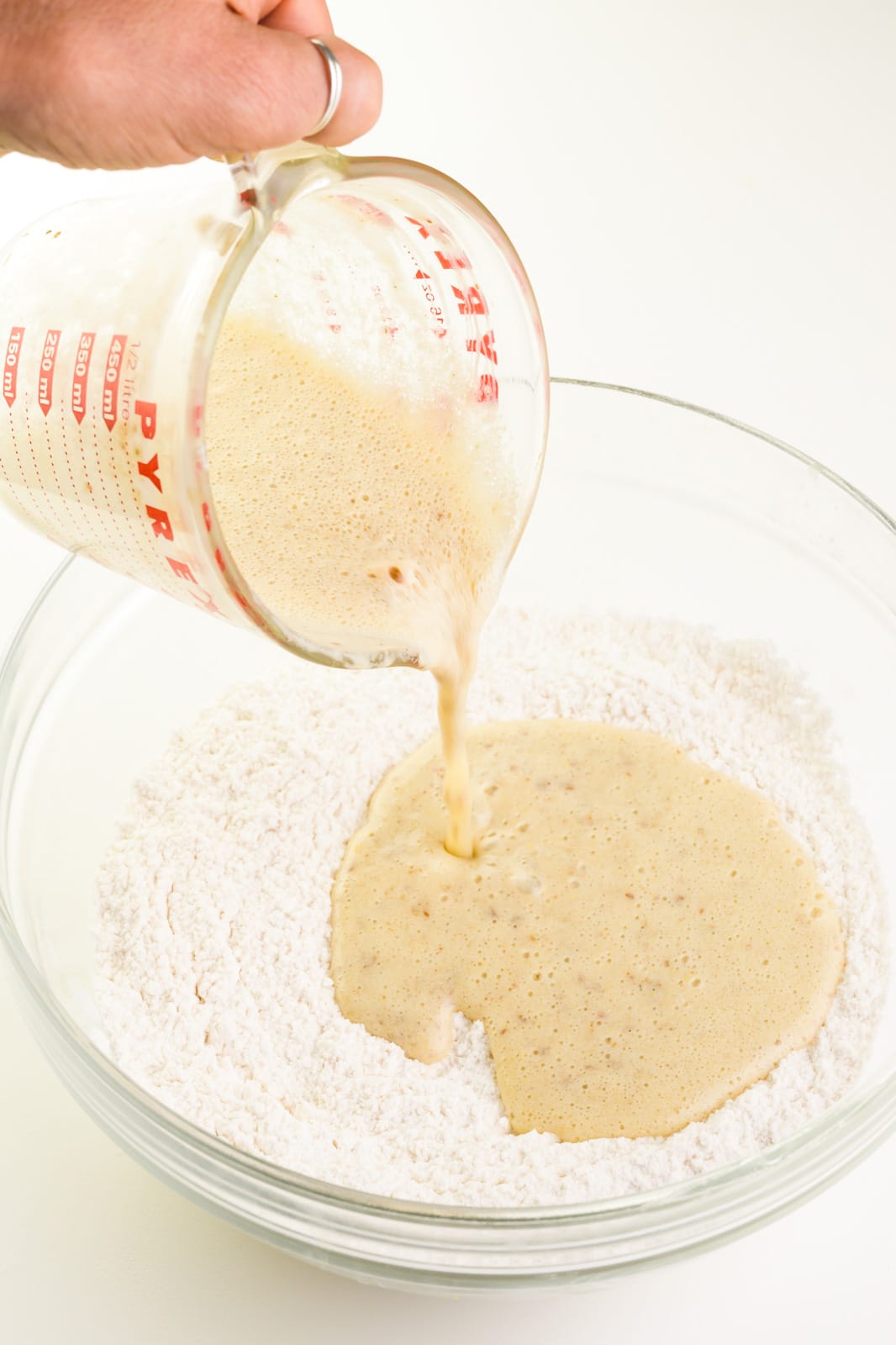 A hand holds a pyrex measuring cup full of a yeast mixture. It's being poured into a flour mixture.