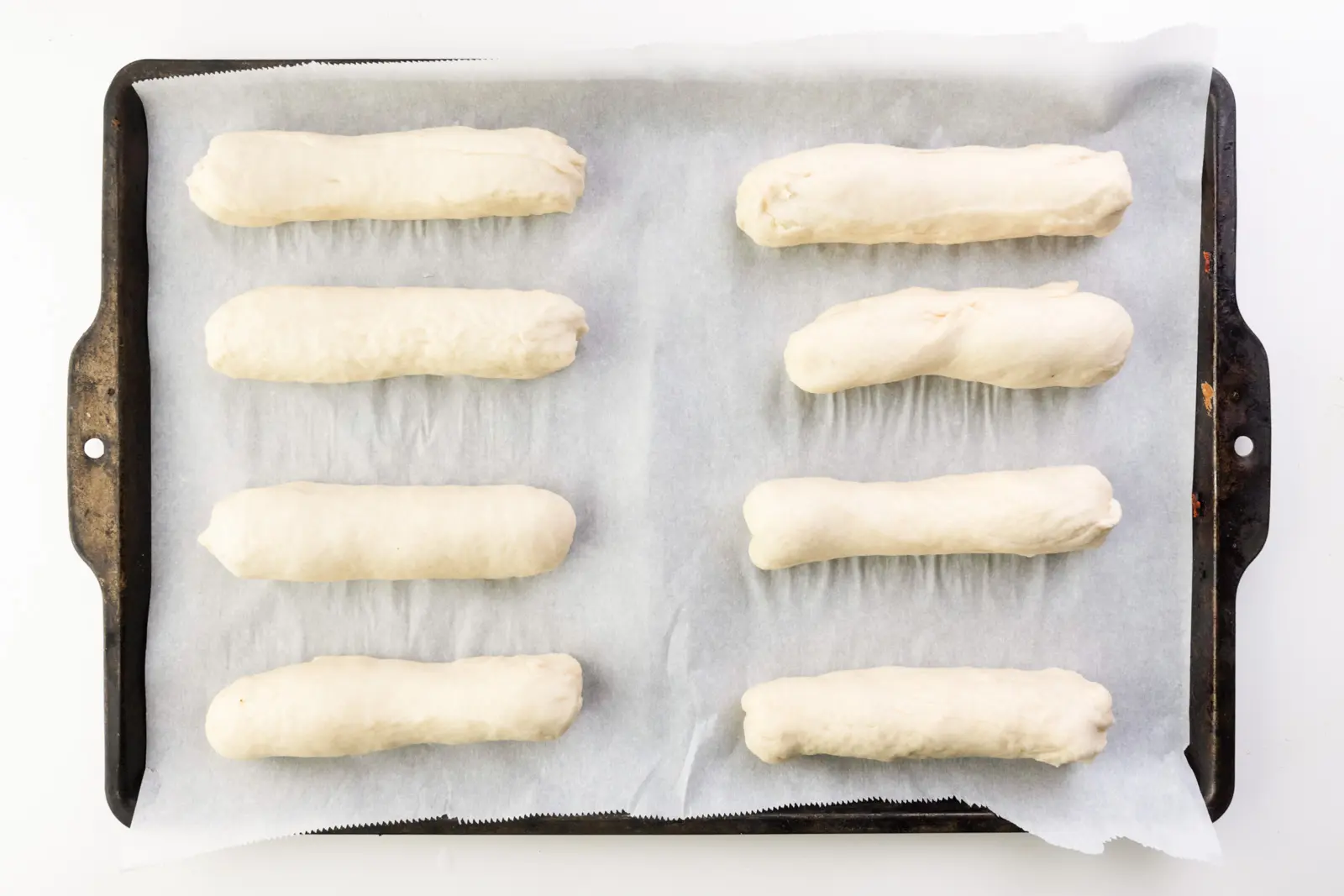 Two rows of unbaked hot dog buns sit on a pan lined with parchment paper.