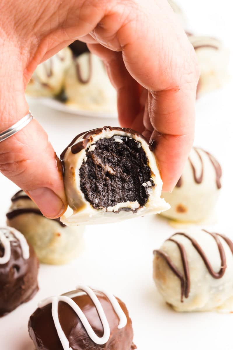 A hand holds an Oreo truffle with a bite taken out. There are more truffles around them, some with dark chocolate coating others with white chocolate coating.